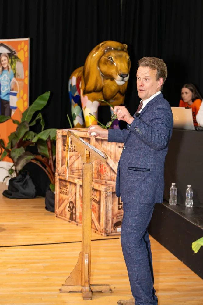 A man in a blue checked suit speaks from a wooden rostrum on stage, with a multicoloured lion sculpture behind him and staff at laptops to the right.